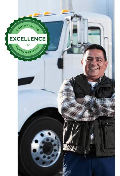 Man smiling with arms crossed while standing beside a large white semi-truck in bright outdoor light.