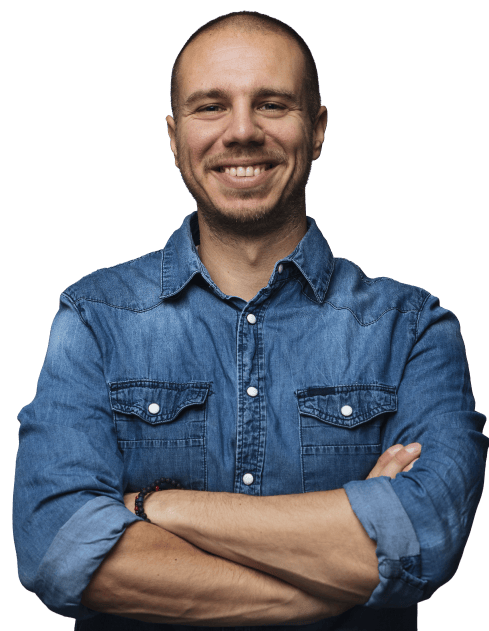 Man smiling with arms crossed while wearing a denim shirt against a simple light grey background.